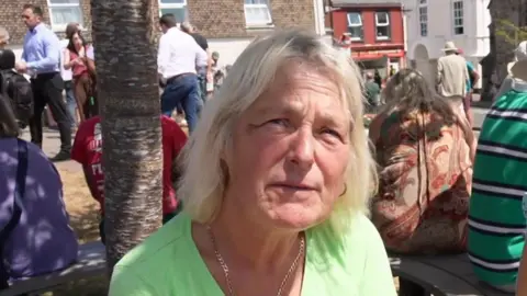 Jacqueline Jones. She has medium length blonde hair and is wearing a bright green top. She is sitting on a bench with other people around a tree.