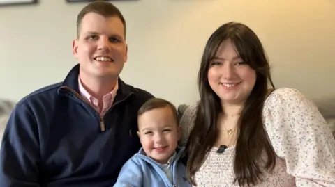 Alex Dunlop/BBC Alaric sits with his mother and father either side of him. All three are smiling broadly. 