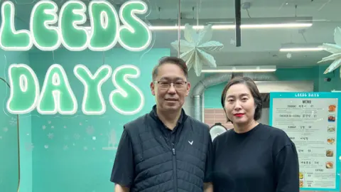 Hyung Joon Kim A middle-aged couple stand outside a glass-fronted restaurant bearing the name 'Leeds Days'. A menu can be seen behind them.