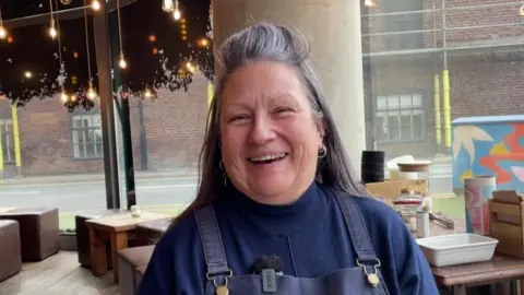 Sofía Luis-Hobbs/BBC Woman in navy blue turtleneck jumper and a matching pinafore dress layered on top smiles at camera. There is a string of fairy lights hanging from the ceiling behind her, and there are wooden tables and stalls in the background. There is a panel of floor-to-ceiling windows behind her which reveals scaffolding on the street outside.