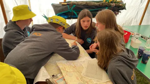 A group of five children is sitting around a table with maps spread out in front of them. They are talking to each other and taking notes.