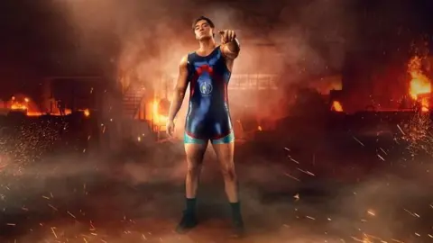 Apollo from Gladiators. He has short brunette hair and is wearing a blue body suit with red and silver detailing. He is holding his chin up and pointing towards the camera. The image has been edited to include fire in the background