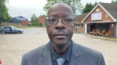 A head shot of Maxwell Muchenje. He is a black man wearing glassed and a grey suit, tie and shirt. Behind him is a car park.