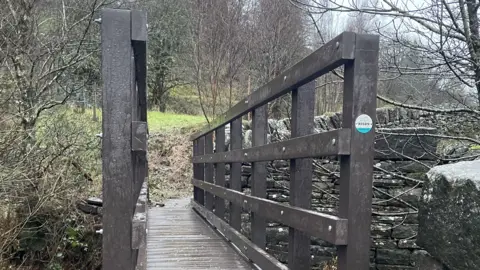 A view across a small footbridge, leading to an area with grass and trees. There is a drystone wall to the right, and a small disc highlighting a walking route is on the righthand pillar of the bridge.