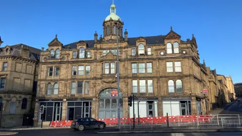 A large historic stone building with arched windows, ornate detailing and a central dome stands on a sunny day. Red plastic barriers line the pavement in front, and a black car is parked on the street. 