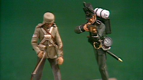 Closeup of two model soldiers with a green background behind them.  Solder on the left is standing with arms by side holding top of his rifle dressed in light green uniform.  Soldier on right looks like a rifelman from the Napoleonic wars, with dark uniform, including tall hat with feather and a backpack, poised in a shooting position.