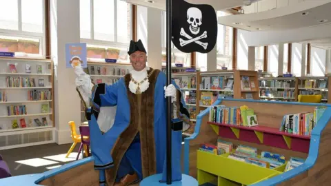A man dressed as a pirate holding a purple book. He is wearing a brown and blue outfit with a black hat is and also holding a skull and crossbones sign on a black pole. He is standing in a library with books on shelves surrounding him.