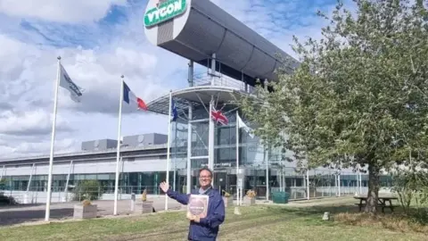 Ajay Chowdhury Ajay Chowdhury wearing a blue jacket and holding his book in one arm with the other arm outstretched, standing in front the building - a futuristic looking glass-panelled building with flags in the foreground and Vygon written in white writing on a green lozenge.