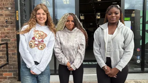 Three teenage girls stand in a line with their hands clasped in front of them outside the front entrance of a building.