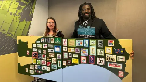Miranda van Rossum and Kofi Smiles holding the embroidery it has a black outline of Hull next to the blue of the Humber Estuary with brightly coloured stiches squares arranged across it.