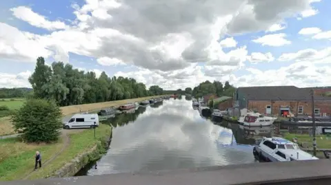 A canal, pictured from a bridge above. Trees and industrial buildings can be seen flanking the water.