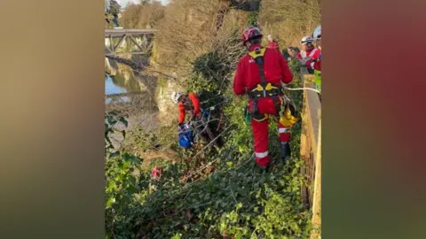 Handout Image shows the rescue team at work, dressed in red outfits and wearing harnesses and hard hates. Three team members peer over a wall at the cliff's edge, while two others in harnesses attached to rope look down the cliff to an unseen sixth team member pulling Bracken up. The cliff is covered with greenery and a bridge can been seen in the background. 