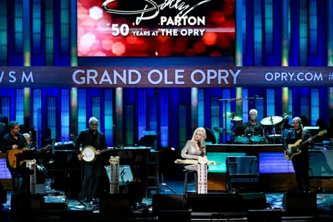 Katherine Bomboy/NBC/NBCU Photo Bank via Getty Images Dolly Parton sits on stage at Opry show. The backdrop is blue and red with a large screen saying it celebrates her 50 years at the Opry. A small band is on stage with her. 