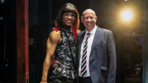 Steve Pateman standing next to Johannes Radebe. Steve is wearing a blue jacket, white shirt and tie, Johannes, in a long wig, and a black and white dress. They are both standing on a theatrical stage and smiling and looking at the camera. A light is on behind them.