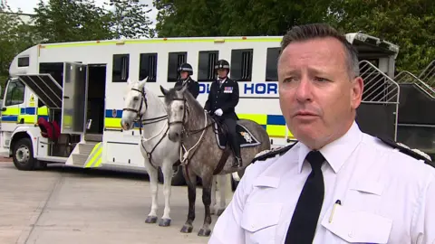 A man in a white uniform standing in front of a police horse transport vehicle, with two mounted officers and horses in the background.