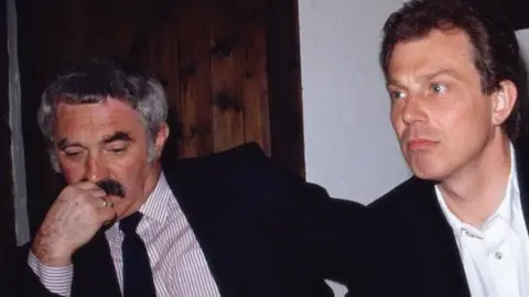 John Burton and Tony Blair are sitting side-by-side at a table. On the left, Burton, who has short greying hair and a thick black moustache, has his hand up to his mouth. He is wearing a dark suit and a striped shirt and tie. Blair, on his right, has short brown hair and is wearing a black suit and white shirt.