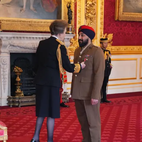 Yui Mok/PA Wire Princess Anne places a medal on Captain Jay Singh-Sohal's jacket in a room at St James's Palace. He wears light brown military uniform and a dark blue and red turban. Princess Anne wears a dark skirt suit with gold embellishments. 