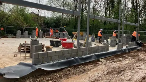 Tom Ingall/BBC Several construction workers in orange safety clothes and white hard hats are building a structure from concrete blocks. Steel beams rise from the foundation, with tools, materials and fencing surrounding the busy site.