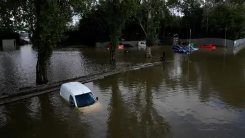 A white van is seen half covered by water in what appears to be a car park. Trees and a compound with vehicles are visible in the background.