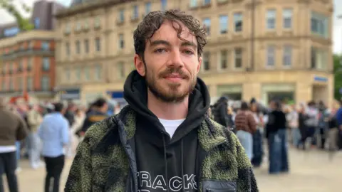 A man stands in a square, surrounded by people. He is wearing a black hoodie and a fleece coat.
