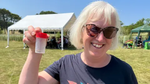 Elaine is smiling, she is wearing a black top and brown lensed sunglasses. She is holding up a transparent plastic container with a red lid which contains a brown bug.