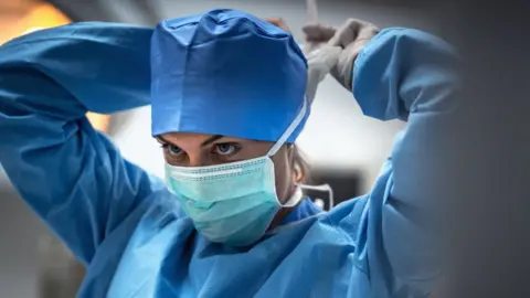 A female health worker tying a face mask on, she is wearing blue scrubs and a hat