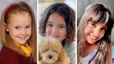  Elsie, who has long fair hair and is wearing a burgundy jumper and a yellow shirt as she smiles for a studio portrait; Alice who has long dark hair is wearing a white top and Bebe is holding a dog with curly dark hair and is wearing a grey top and dark trousers. 