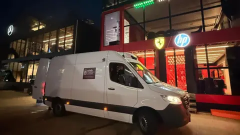 Towcester Community Larder A white van is parked at Silverstone outside two buildings, one displaying a lighted Mercedes sign and the other a lighted Ferrari sign.