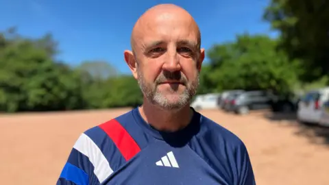A man stood in a large car park. He is bald, has a grey and black beard and is wearing a blue Adidas sports top with red blue and white stripes on his shoulder. There are cars parked on a red gravel car park behind him.