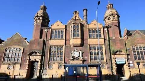 A large historic building which has pointed roof accents and lots of large windows. Above two dark blue doors is a stone sign which reads "women's baths" and there are two stone people next to a large crest.