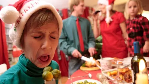 A young boy wearing a festive hat grimaces as a fork full of brussels sprouts approaches his mouth. Behind him, his family cheerfully get on with Christmas dinner.