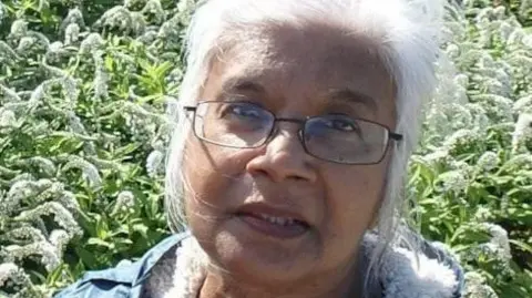 A family handout of Mena. She has long white hair and is wearing glasses. She is standing in front of a garden.