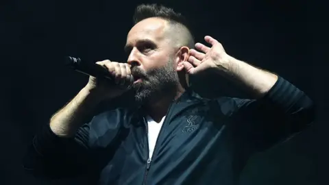PA Media Head and shoulders image of Alfie Boe on a black stage singing. He is holding a mic in his right hand and had his left hand behind his ear as though he is listening. He is wearing a black tracksuit top over a white tshirt and his hair is shaved at the sides, longer on top and he has a beard with flecks of grey