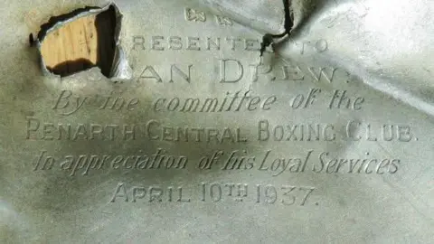 A buckled silver cigarette case: the top right is folded in upon itself, whilst in the top left a bullet hole obscures the inscription