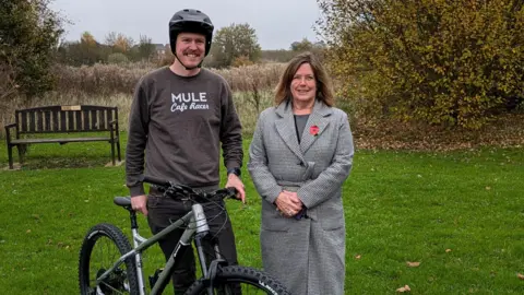 Olly Rose A man and woman stand next to each other on grass in front of a wooden bench. The man is holding up a black bike, wearing a black bike helmet, dark grey jumper and black jeans. The woman has shoulder-length brown hair and is wearing a long black and white checked coat with a poppy at the lapel. Long grass and shrubs can be seen in the background.