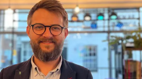 Alex Hughes is standing inside the community hub. He has a beard and circle-framed glasses and is wearing a navy blazer and a light blue shirt. He is smiling at the camera