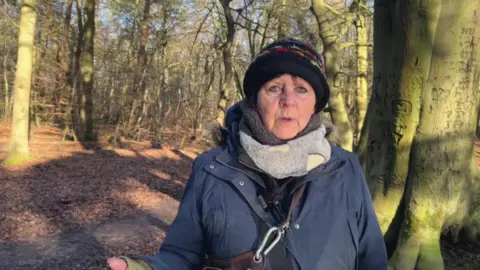 BBC/Nathan Turvey Commercial dog walker Linda Riding wearing a hat, scarf and coat with trees behind her on a sunny day.