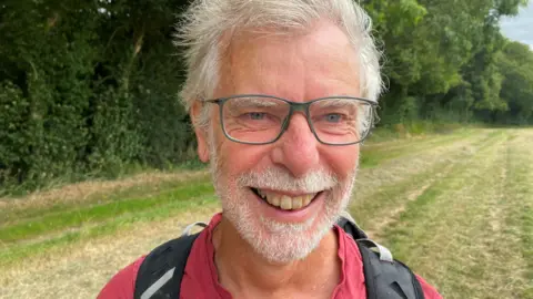 Emily Unia/BBC An older man with white hair and beard and glasses smiling at the camera, with a field and trees in the background