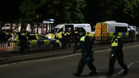 Getty Images Two police officers walking across the road, with more in the background standing beside a police car and two vans. 