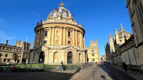 It's a sunny day in Oxford. A big domed buiding is to the fromt with historic buildings around it. Bicycles are chained on the fence around the central building.