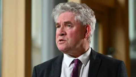 Getty Images Kenneth Gibson, a man with curly grey hair, photographed in the Scottish Parliament. Visible from the shoulders up, he is wearing a black suit, white shirt and purple tie. 