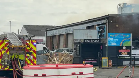 Stephen Huntley/BBC The back of a fire engine parked by the side of the road. It is parked by an industrial site and there is smoke in the air coming off from one of the buildings which is on fire.