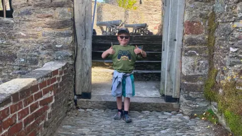 Contributed A young boy stands in an archway smiling at the camera with his thumbs up. He is wearing a cap, sunglasses, a green T-shirt and shorts with a blue top tied around his waist.