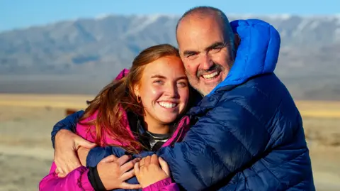 Andrew with his arms wrapped around Molly. Andrew is bald, wearing a blue puffer coat. Molly has red hair tied back and is wearing a pink puffer coat.