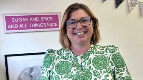 Kate Bradbrook/BBC Woman in a green and white patterned dress with short light brown hair and glasses stands in front of a pink sign which reads "Sugar and spice and all things nice"