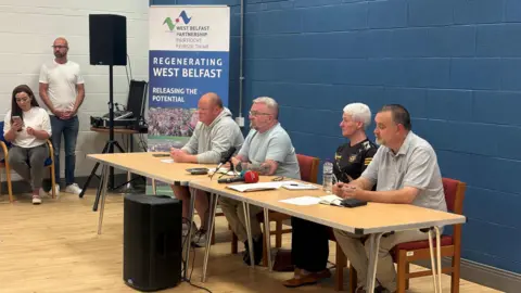 A tale with four people sat at it. On the table, there are a number of microphones, including a read microphone. The wall behind the table is dark-blue. On the left of the tale is a large sign which reads: 'REGENERATING WEST BELFAST - RELEASING THE POTENTIAL'. There is a man stood next t the sign, and a woman sat next to him. 