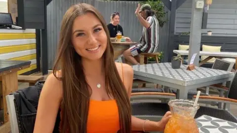 Family handout A woman with long brown hair and wearing an orange dress. She is sitting outside a bar with an orange drink. 