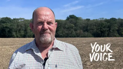 Will Fenn is looking at the camera and standing in a field with trees in the distance. He is balding with facial hair and is wearing a checked shirt.