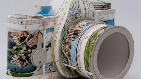 Jenifer Collier A piece of artwork in the shape of an SLR camera, made out of paper and featuring prints of mountains on it