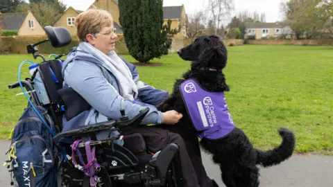 Canine Partners Samantha with short hair. She is sitting on a motorised wheelchair with her black dog standing on its two legs looking at her face.